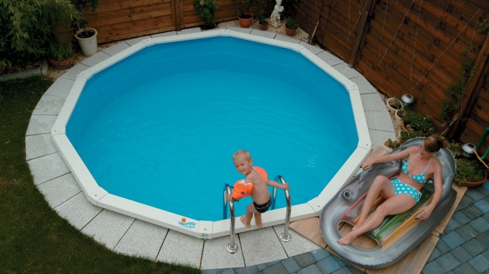 Cranpool Riva ist der kleine Pool für jeden Garten. Am Bild ist ein rundes Schwimmbecken Riva. Im Vordergrund liegt eine junge Dame auf einem Liegestuhl. Ein kleiner Junge steht an der Poolleiter in Badehose und Schwimmflügel und ist für den Sprung ins Schwimmbecken bereit. Hinter dem Pool befindet sich Holzelemente als Sichtschutz. Das Pool Riva ist in die Erde eingebaut und ist mit hellen Pflastersteinen umrandet.