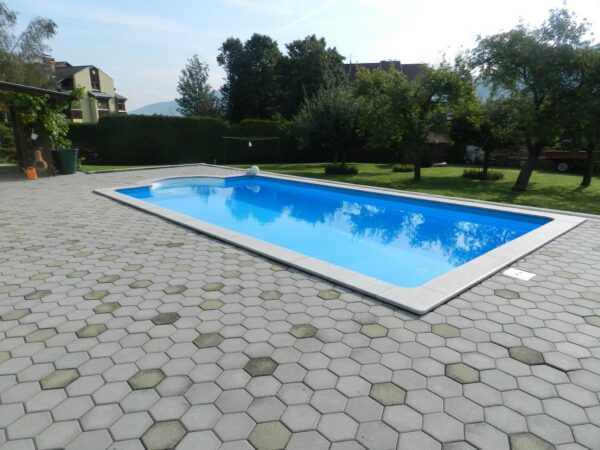 Cranpool Cranthermo ist ein selbstgemauertes Rechteckpool. Auf dem Bild zu sehen ist ein Schwimmbecken Cranthermo in einem schönen großen Garten. Das Schwimmbecken ist von grauen bienenwaben förmigen Pflastersteinen umgeben. Im Hintergrund sind mehrere Obstbäume und eine Hecke zu sehen.