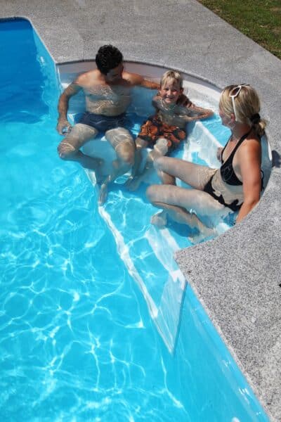 Heimwerker-Schwimmbecken Cranthermo ist ein Rechteckpool für Heimwerker. Das Bild zeigt einen Mann eine Frau und ein Kind auf der Einstiegstreppe sitzend.
