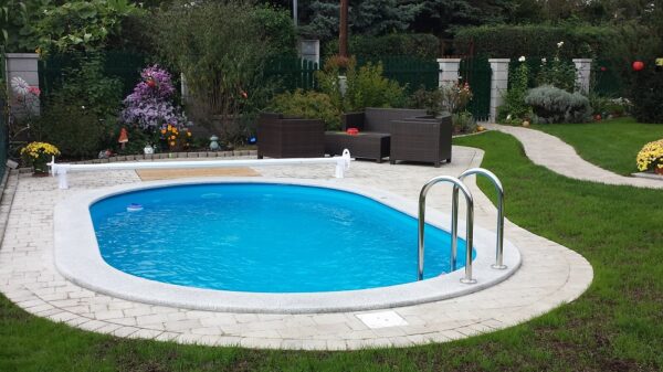 Einbau-Schwimmbecken Miami ist für Ästheten und Individualisten konzepiert. Das Bild zeigt ein ovales Schwimmbecken Miami. Der Beckenrand ist aus Granit. Rund um den Beckenrand wurden helle Pflastersteine verlegt. Im Hintergrund ist ein schön angelegter Garten zu sehen. Am Beckenrand steht eine Sitzgarnitur. Für die Abdeckung den Schwimmbecken ist eine Abdeckplane vorgesehen.