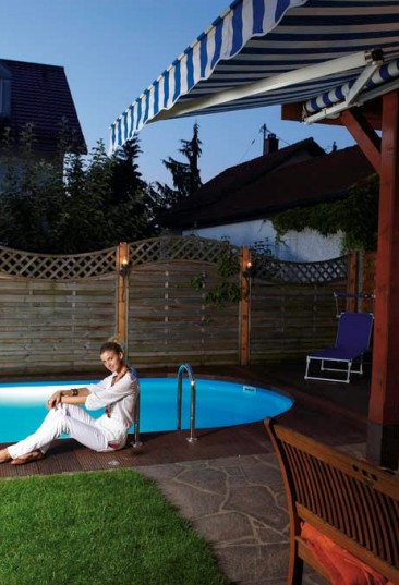 Einbau-Schwimmbecken Miami bietet ein auf die Haus-und Gartenarchitektur abgestimmtes Schwimmparadies. Auf dem Bild sieht man ein ovales kleines Schwimmbecken Miami. Der Beckenrand ist mit Lärchenbrettern verbaut. Auf dem Beckenrand sitzt eine Frau in weißer Bekleidung. Auf der Terrasse vor dem Schwimmbecken stehen ein Tisch und eine Bank. Der Sichtschutz im Hintergrund ist durch Holzelemente gegeben.