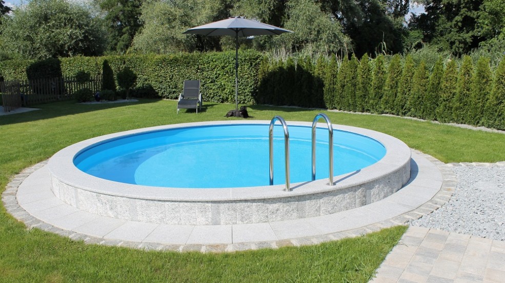 Einbau-Schwimmbecken Miami lässt viele Gestaltungsmöglichkeiten zu. Das Bild zeigt ein rundes Schwimmbecken Miami. Der Beckenrand wurde aus grauen Steinen gefertigt. Am Beckenrand wurde ein Kreis aus hellen Steinen verlegt. Der Sichtschutz zum Schwimmbecken wird durch eine Hecke gegeben. Im Hintergrund steht ein Liegestuhl mit aufgespannten Schirm darunter liegt ein schwarzer Hund.