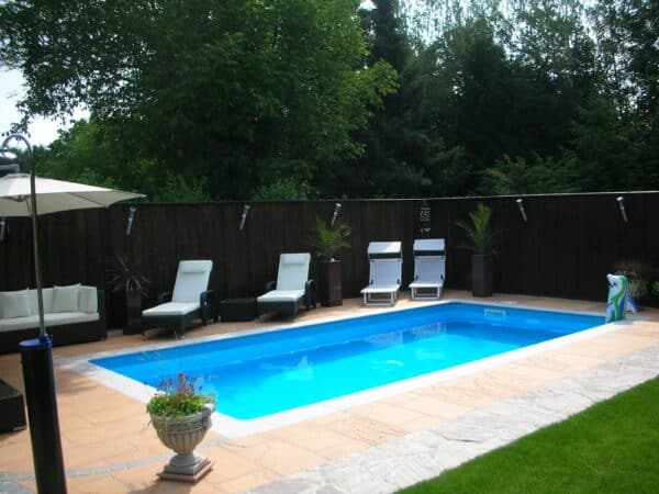 Cranpool Ozean besticht durch eine rasche Bauzeit. Das Bild zeigt ein Schwimmbecken Ozean. Rund um den Beckenrand wurden helle Pflastersteine verlegt. Der Sichtschutz im Hintergrund ist durch eine Holzwand gegeben. Vor dieser Holzwand stehen vier Liegestühle und eine Bank mit weißen Bezug. Am linken Beckenrand steht eine Solardusche.