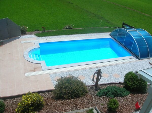 Ozean ist ein Rechteck-Schwimmbecken das zeitlosen Stil mit langlebiger Schönheit vereint. Am Bild sieht man ein Schwimmbecken Ozean mit einem geöffneten Cabriodom. Auf der linken Breitseite befindet sich die Römertreppe. Vor der Treppe wurden helle Platten verlegt. Auf den Längsseiten des Schwimmbeckens befinden sich die Schienen für den Cabriodom. Vor den Schienen sind weiße Kieselsteine. Im Vordergrund des Bildes sieht man noch ein paar Stauden. Im Hintergrund ist eine schöne Wiese.