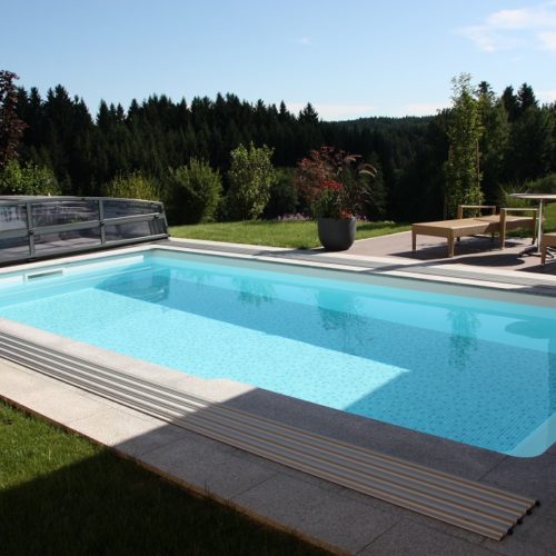 Das Rechteck-Schwimmbecken Ozean vereint zeitlosen Stil mit langlebiger Schönheit. Das Bild zeigt ein Schwimmbecken Ozean mit geöffneten Cabriodom. Im Hintergrund ist ein Wald und Sräucher zu sehen. Am Beckenrand steht ein kleiner Tisch mit zwei Liegestühle und ein geschlossener Schirm.