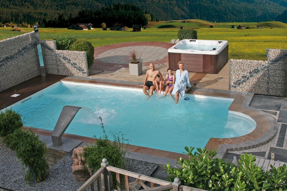 Cranpool Ozean besticht durch eine rasche Bauzeit. Das Bild zeigt ein Schwimmbecken Ozean mit integrierter Römertreppe. Das Schwimmbecken ist von roten und grauen Pflastersteinen umgeben. Am Beckenrand sitzt eine Frau, zwei Kinder und ein Mann. Hinter ihnen steht ein Whirlpool. Im Hintergrund ist eine große Wiese, Wald und mehrere Hütten zu sehen. Der Sichtschutz wird links und rechts vom Schwimmbecken durch Steinmauern gebildet. Im Vordergrund sind mehrere Grünpflanzen zu sehen.