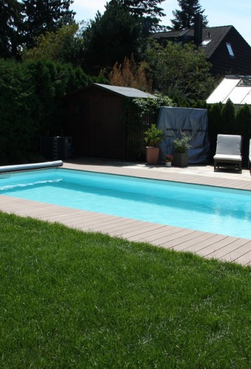 Rechteck-Schwimmbecken Ozean ist das High-Class-Schwimmbecken made in Austria. Das Bild zeigt ein Schwimmbecken Ozean das von hellen Holzbrettern umgeben ist. Im Hintergrund sind Häuser zu sehen. Der Sichtschutz ist durch eine Hecke gegeben. Am Beckenrand sind zwei Liegestühle mit weißer Auflage aufgestellt.