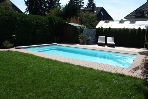 Rechteck-Schwimmbecken Ozean ist das High-Class-Schwimmbecken made in Austria. Das Bild zeigt ein Schwimmbecken Ozean das von hellen Holzbrettern umgeben ist. Im Hintergrund sind Häuser zu sehen. Der Sichtschutz ist durch eine Hecke gegeben. Am Beckenrand sind zwei Liegestühle mit weißer Auflage aufgestellt.
