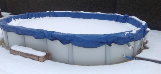 Cranpool PE-Winterabdeckplane schützt Ihr Schwimmbecken in der kalten Jahreszeit. Das Bild zeigt ein ovales Schwimmbecken Sun Remo mit einer blauen Winterabdeckung. Im Hintergrund ist eine Hecke und Häuser zu sehen. Das Becken ist von Schnee bedeckt und umgeben.