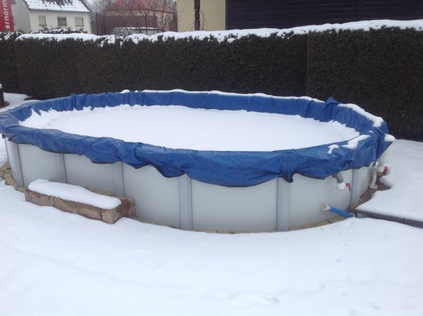 Cranpool PE-Winterabdeckplane schützt Ihr Schwimmbecken in der kalten Jahreszeit. Das Bild zeigt ein ovales Schwimmbecken Sun Remo mit einer blauen Winterabdeckung. Im Hintergrund ist eine Hecke und Häuser zu sehen. Das Becken ist von Schnee bedeckt und umgeben.
