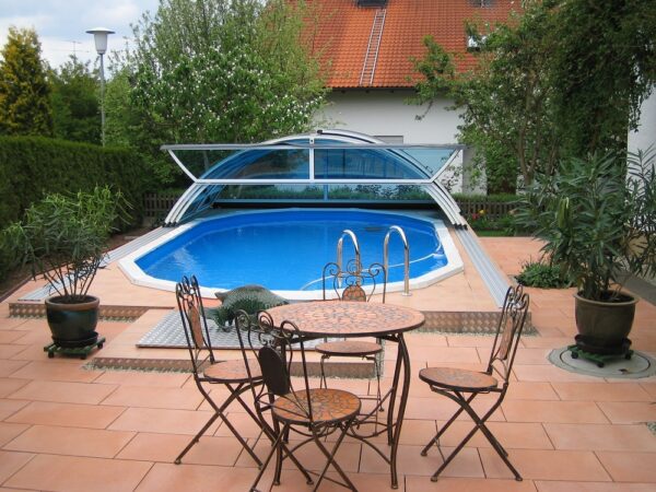 Stahlwand-Swimmingpool Royal wird in Österreich hergestellt. Das Bild zeigt ein Stahlwand Schwimmbecken Royal mit geöffneten Cabriodom. Rund um das Becken sind roto Steine verlegt. Im Hintergrund ist ein Familienhaus. Davor sind einige Bäume zu sehen. Vor dem Schwimmbecken steht ein Tisch mit vier Stühlen. Links und rechts vom Becken stehen Grünpflanzen.