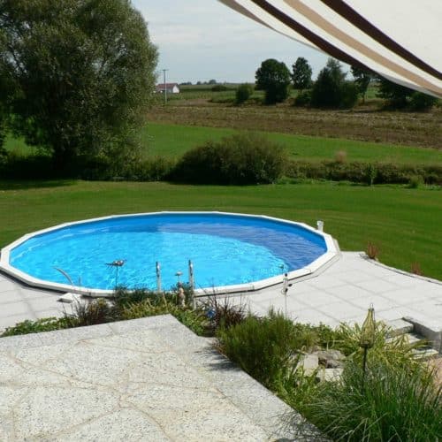 Cranpool Royal besticht durch seine stabile Konstruktion. Das Bild zeigt ein rundes Schwimmbecken Royal das komplett in die Erde eingebaut ist. Im Vordergrund des Beckens wurden graue Steinplatten verlegt. Im Hintergrund ist eine Wiese mit anschließenden Acken.