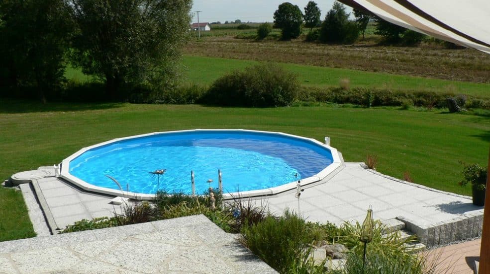 Cranpool Royal besticht durch seine stabile Konstruktion. Das Bild zeigt ein rundes Schwimmbecken Royal das komplett in die Erde eingebaut ist. Im Vordergrund des Beckens wurden graue Steinplatten verlegt. Im Hintergrund ist eine Wiese mit anschließenden Acken.