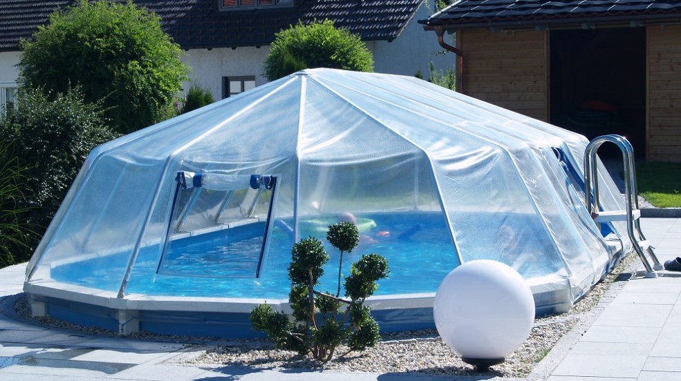 Cranpool Sonnedom ist eine preisgünstige Überdachung. Das Bild zeigt ein Schwimmbecken Royal mit geschlossenen Sonnendom. Der Beckenrand ist mit hellen Pflastersteien umrandet. Im Hintergrund ist ein Einfamilienhaus und eine Gartenhütte. Vor dem Becken ist eine Grünpflanze und eine runde Beleuchtung.