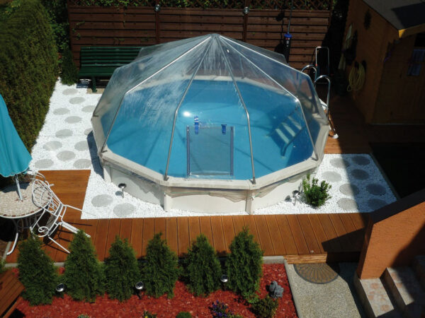 Cranpool Sonnendom ist für runde und ovale Becken erhältlich. Das Bild zeigt ein Schwimmbecken Riva mit Sonnendom in einem kleinen Garten. Das Schwimmbecken ist von Lärchenbrettern und hellen Steinen umgeben. Im Hintergrund bilden Holzelemente und links vom Schwimmbecken Thujenhecken einen Sichtschutz.