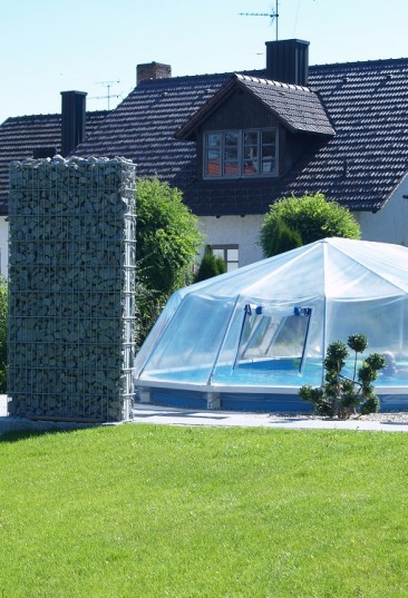 Cranpool Sonnendom sorgt für höhere Wassertemperaturen. Das Bild zeigt einen geschlossenen Sonnendom. Im Hintergrund befindet sich ein Einfamilienhaus. Das Schwimmbecken ist in die Erde eingebaut und von hellen Steinen umgeben. Am linken Beckenrand stehen zwei Steinsäulen.