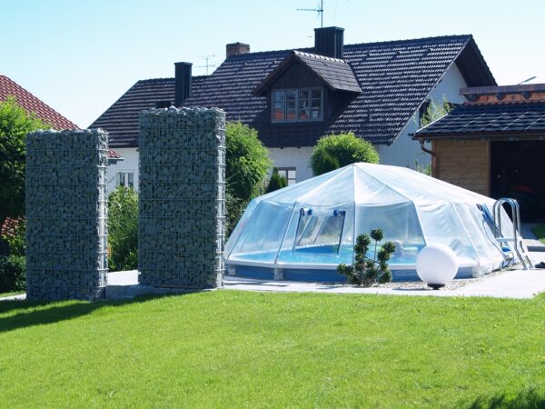 Cranpool Sonnendom sorgt für höhere Wassertemperaturen. Das Bild zeigt einen geschlossenen Sonnendom. Im Hintergrund befindet sich ein Einfamilienhaus. Das Schwimmbecken ist in die Erde eingebaut und von hellen Steinen umgeben. Am linken Beckenrand stehen zwei Steinsäulen.