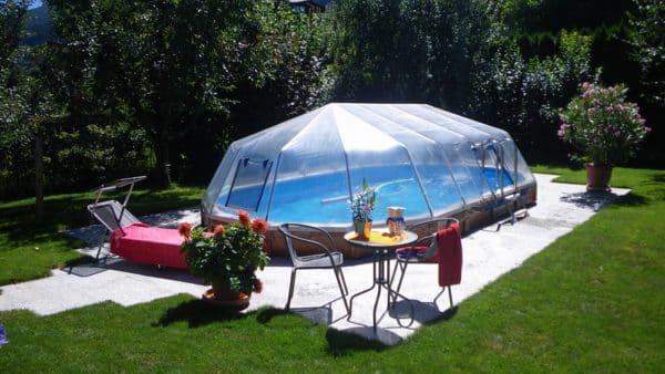 Cranpool Ovales Becken mit einem Sonnendom. Das Schwimmbecken ansich ist mit Holz verkleidet. Der Sonnendom hat zwei Einstiege, wobei bei eiem eine dreistufige Leiter steht, Vor dem Schwimmbad erknnt man einen Kaffetisch mit zwei Sttühlen und eine Sonnenliege. auf der Sonnenliege liegt ein Hadtuch. Im Hintergrund erkennt man einige Bäume und einen Rasen in einem Garten. Das Schwimmbecken ansich ist mit grauen Steinen umgeben.