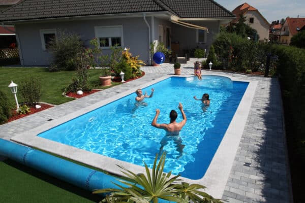 Cranpool Ihr Spezialist für Schwimmbecken im rechteckigen Schwimmbad befinden sich drei Personen, die Wasserball spielen. Auf der halbrunden vierstufigen Treppe sitzt eine Frau und sieht dem Geschehen zu. Das Schwimmbecken ist von Pflastersteinen umgeben. Auch kann manim garten eine Abdeckplane erkennen.