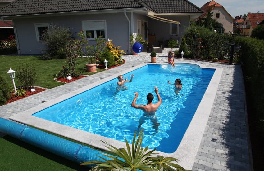 Cranpool Ihr Spezialist für Schwimmbecken im rechteckigen Schwimmbad befinden sich drei Personen, die Wasserball spielen. Auf der halbrunden vierstufigen Treppe sitzt eine Frau und sieht dem Geschehen zu. Das Schwimmbecken ist von Pflastersteinen umgeben. Auch kann manim garten eine Abdeckplane erkennen.