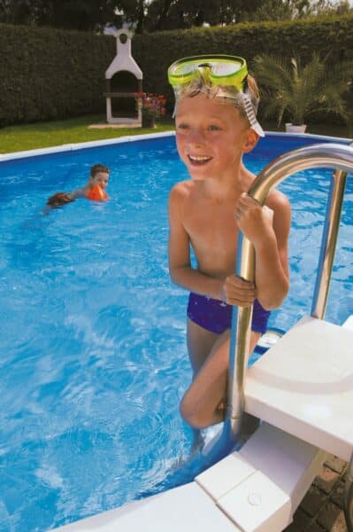 Stahlwand-Swimmingpool Royal ist die stabilste Konstruktion für runde und ovale Becken am Markt. Das Bild zeigt einen kleinen Jungen der auf der Eistiegstreppe steht und eine Taucherbrille am Kopf hat. Im Hintergrund schwimmt ein kleiner Junge im ovalen Becken Royal. Einen Hecke bildet den Sichtschutz zum Schwimmbecken. Vor der Hecke steht ein Griller.