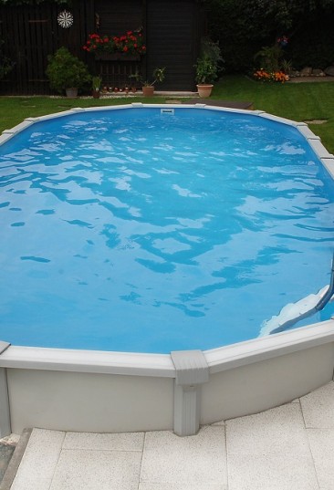 ein ovales Sun Remo Schwimmbecken von Cranppool in einem Garten. Das Pool ist oval und von weißen und grauen Pflastersteinen umgeben.
