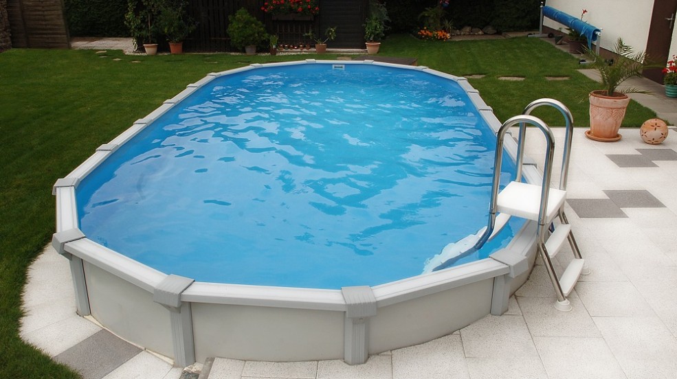 ein ovales Sun Remo Schwimmbecken von Cranppool in einem Garten. Das Pool ist oval und von weißen und grauen Pflastersteinen umgeben.