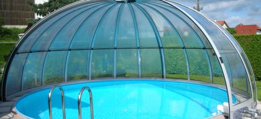 Cranpool Rundhallen sind eine ideale Lösung für eine Ganzjahresabdeckung von Rundbecken. Das Bild zeigt ein rundes Schwimmbecken mit einer Rundhalle die geöffnet ist. Der Beckenrand ist mit hellen Pflastersteinen umrandet. Im Hintergrund sieht man einige Häuser und eine Fabrikshalle. Der Sichtschutz zum Schwimmbecken wird mit einer Hecke gebildet.