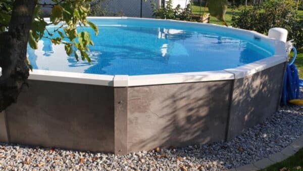 Ovales Schwimmbecken von Cranpool mit grauer Holzverkleidung und einer Kieselstein umrundung in einem Garten