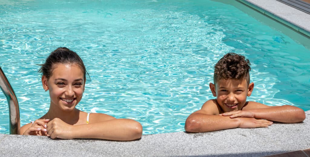 Frau und Bursche im Wasser lehnen am Beckenrand und lächeln im Hintergrund Pool