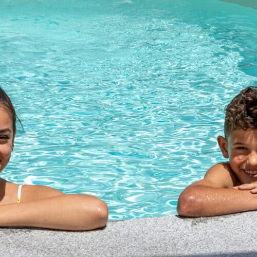 Frau und Bursche im Wasser lehnen am Beckenrand und lächeln im Hintergrund Pool