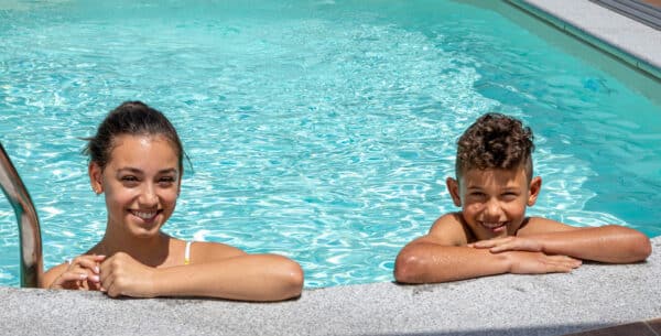 Frau und Bursche im Wasser lehnen am Beckenrand und lächeln im Hintergrund Pool