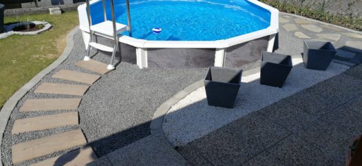 Rundbecken Riva mit einer grauen Verkleidung und einem Steinumfeld mit Kies und stein pplatten. Surch den Kies führt ein Weg zum SChwimmbad