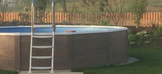 rundbecken mit hellbrauner Verkleidung in einem Garten. Zu dem Pool führt ein Weg aus Steinen durch das Gras. Im Hintergrund erkennt man Bäume und andere Pflanzen in einem beet.