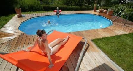 ein eingegrabenes oval becken mit einem Holzdeck und einem Holzsteg rund ums Pool. IM Cranpool Schwimmbecken sind zwei Kinder, während die Mutter auf einer orangen Liege liegt