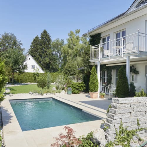 Pool im Garten, das rechteckige Schwimmbecken von Cranpool ist eingegraben und somit auf einer Ebene mit der Terasse . Die Terasse wurde so verlängert, dass rund ums Pool ein Steg gebaut wurde. Im hintergreund erkennt man einen Garten von einem Einfamilien Haus