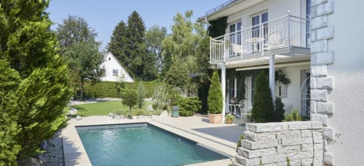 Pool im Garten, das rechteckige Schwimmbecken von Cranpool ist eingegraben und somit auf einer Ebene mit der Terasse . Die Terasse wurde so verlängert, dass rund ums Pool ein Steg gebaut wurde. Im hintergreund erkennt man einen Garten von einem Einfamilien Haus