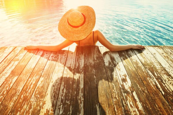 Woman in hat relaxing at the pool eine Frau im Sonnenhut sitzt bei Sonnenuntergang in einem SChwimmbecken. Die sonne fällt so, dass ihr schatten auf das Deck fällt.