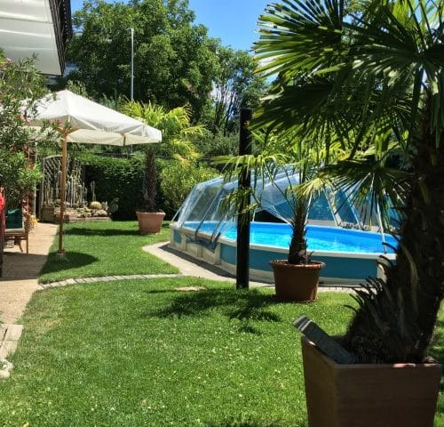 Ein schwimmbecken Royal in einem Garten. Der Pool ist mit einer Sschwimmbadabdeckung abgedeckt. Bei der Überdachung handelt es sich um einen Sonnendom von Cranpool. Der Garten wirkt mit seinem Sonnenschirm und den palmen wie ein Urlaubsparadis.
