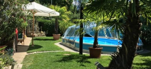 Ein schwimmbecken Royal in einem Garten. Der Pool ist mit einer Sschwimmbadabdeckung abgedeckt. Bei der Überdachung handelt es sich um einen Sonnendom von Cranpool. Der Garten wirkt mit seinem Sonnenschirm und den palmen wie ein Urlaubsparadis.