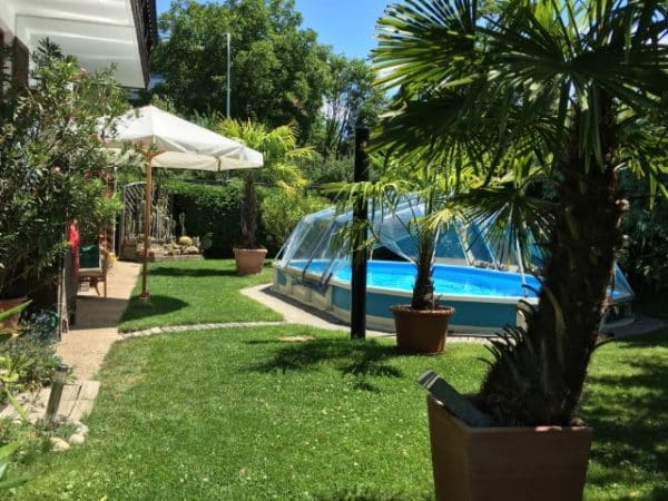 Ein schwimmbecken Royal in einem Garten. Der Pool ist mit einer Sschwimmbadabdeckung abgedeckt. Bei der Überdachung handelt es sich um einen Sonnendom von Cranpool. Der Garten wirkt mit seinem Sonnenschirm und den palmen wie ein Urlaubsparadis.