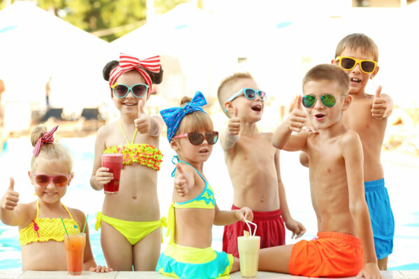 eine Gruppe Kinder, weöche in Badeanzüge, Bikinis und Badehosen einen Tag am Pool genießen, im MOment maxchen sie gerade eine Trinkpasue, alle Kinder haben Sonnenbrillen auf