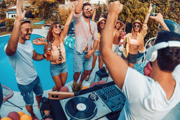 Enjoying pool party with friends.Group of friends having party by swimming pool.eine Gruppe von jungen Erwachsenen bei einer Pool Party, wie sie Musik hören, tanzen und trinken voe einem versenkten ovalen Schwimmbad