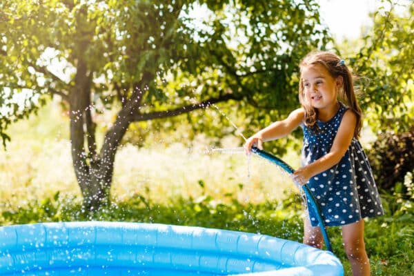 Little girl pouring water into a small pool ein kleines Mädchen, dass ine einem Sommerkleid im Garten versucht das Schwimmbad mit einem Gartenschlauch zu befüllen, dabei geht das Wasser nicht nur ins Pool sondern spritzt auch alles im näöheren Umkreis an