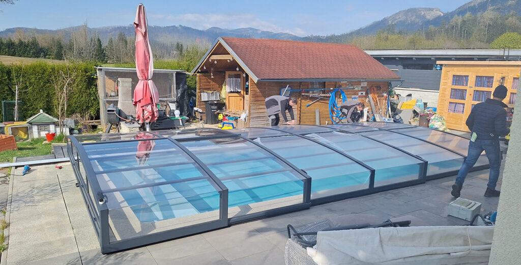 Schwimmbadüberdachung Cabriodom Selekt geschlossen im Hintergrung Holzhütte